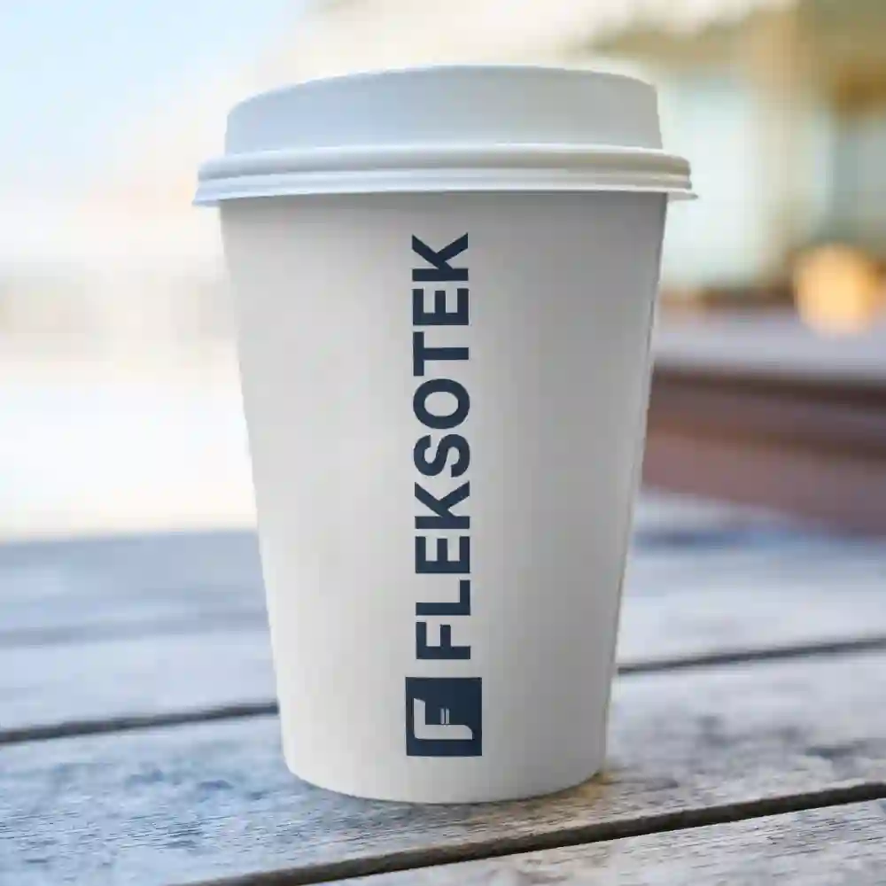 White takeaway coffee cup with vertical Flekstek logo, placed on a wooden table outdoors.